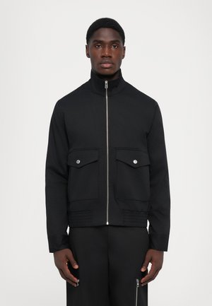 Giacca nera con colletto alto, chiusura frontale con zip, due tasche sul petto con pattine e dettagli texturizzati all'orlo. Tessuto liscio, design aderente.