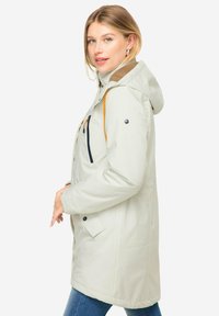 Vaaleanbeige vedenpitävä takki hupulla, jossa on oransseja korostusnyörejä, sivutaskuja ja sileä pinta. Suunniteltu ulkoiluun.