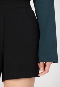 Črne teksturirane hlače s stranskimi žepi, v kombinaciji z dolgimi rokavi v spreminjajoči se barvi teal. Osredotoča se na teksturo tkanine in barvni kontrast.