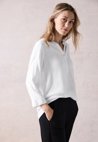 Blusa blanca de manga larga con cuello tipo henley y botones, combinada con pantalones negros. Tejido suave, corte relajado, bolsillo lateral visible.