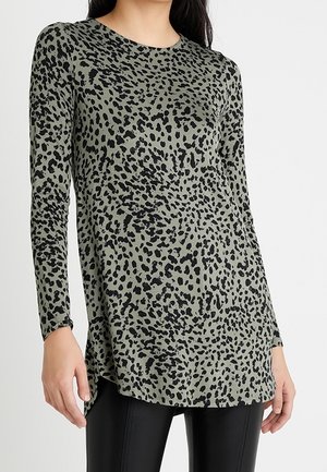 Žena oblečená v olivově zeleném topu s dlouhým rukávem a černým abstraktním leopardím vzorem a černých kalhotách, stojící před jednobarevným pozadím.