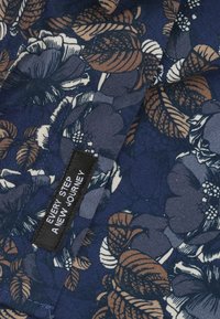 Bloemenstof in marineblauw met grote witte en bruine bloemen en bladeren. Een zwart etiket met de tekst "IEDERE STAP EEN NIEUWE REIS."