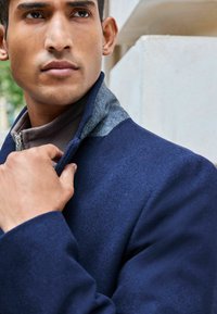 Abrigo azul marino con textura suave, con un cuello gris y cierre de cremallera. La mano del modelo está ajustando el cuello.