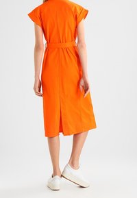 Kvinde iført en lys orange, kortærmet midi-kjole med bælte og slids bagpå, parret med hvide plateausneakers, vendt væk.