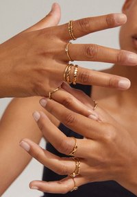 Des bagues en or sur plusieurs doigts, de designs variés : textures tordues, lisses et martelées, avec une petite pierre précieuse transparente sur l'une d'elles.