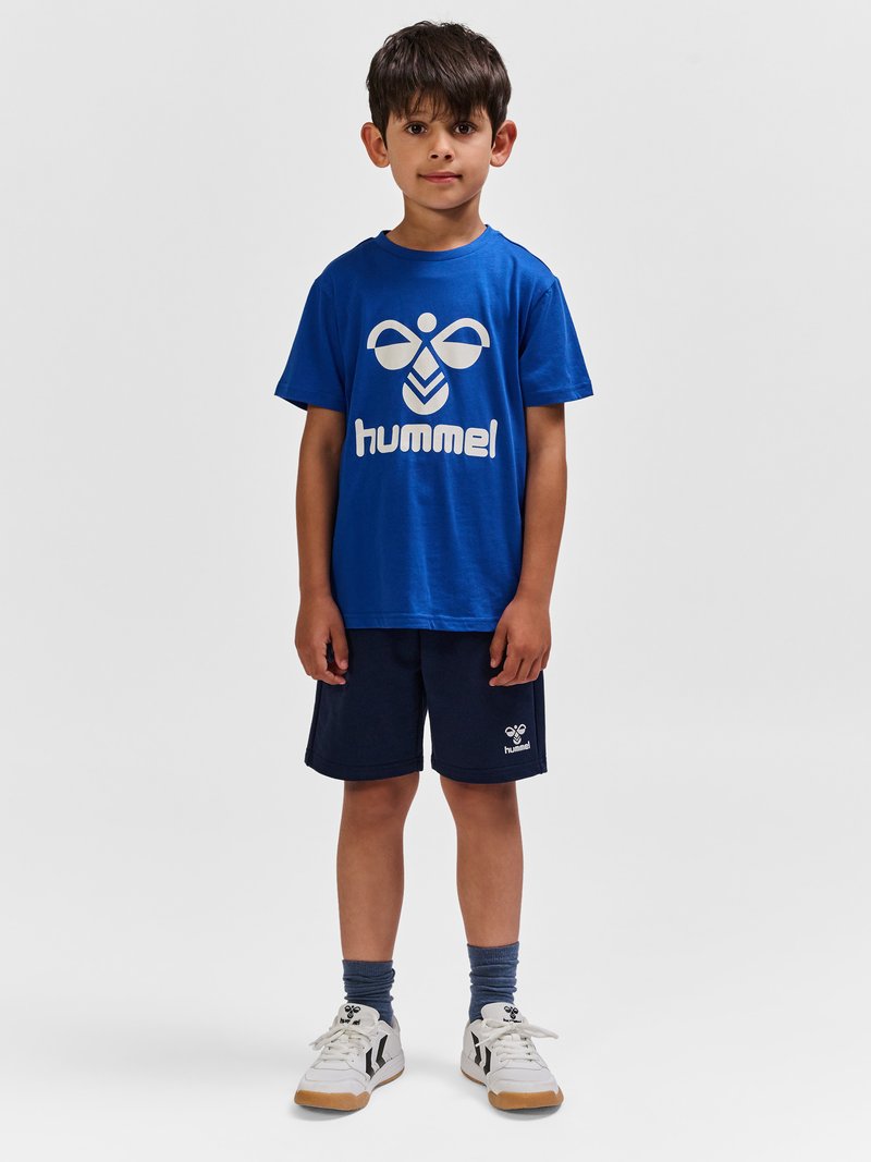 Dreng stående iført en blå hummel t-shirt, marineblå hummel shorts, grå sokker og hvide sneakers på en ensfarvet grå baggrund.