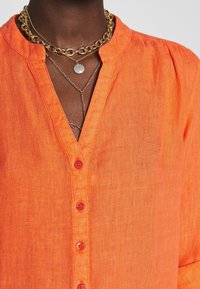 Camicia di lino arancione con scollatura a V, chiusura frontale con bottoni e maniche corte. Accentuata da diverse catene dorate e una collana con pendente di perle.