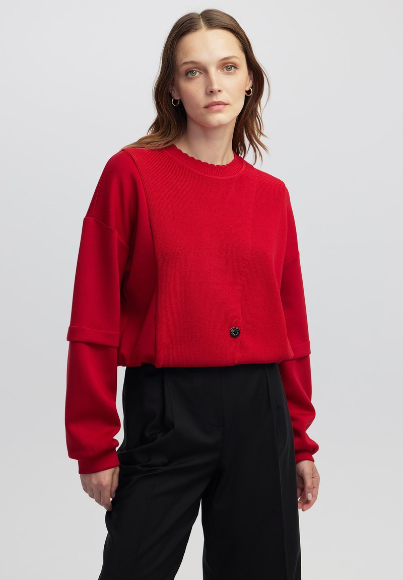 Pull rouge court avec un tricot texturé, un col festonné et un détail de bouton noir rond. Assorti à un pantalon noir taille haute.