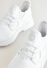 Baskets blanches de sport avec un matériau lisse, un col rembourré, des lacets et un accent de logo texturé au niveau du talon. Design à bout rond.