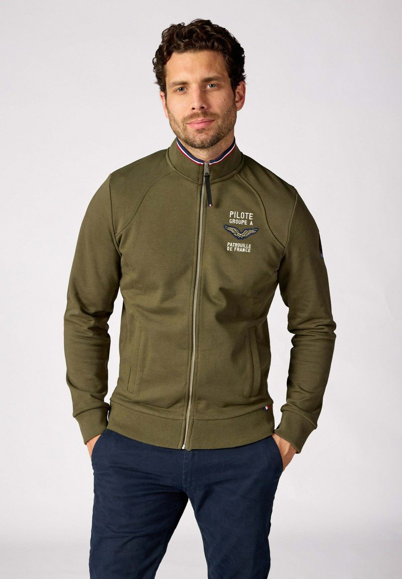 Veste zippée vert olive avec un col montant, ornée d'un logo et d'un design d'aile sur la poitrine, des poignets côtelés et des poches latérales.