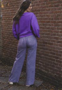 Paarse sweatshirt met een geribde onderrand, gecombineerd met lichtpaarse wijde jeans. De achtergrond toont een gestructureerde bakstenen muur.