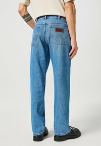Jeans en denim bleu avec une coupe droite, présentant une étiquette en cuir marron à la taille arrière et une couture classique des poches arrière.