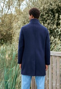 Cappotto in misto lana navy con colletto alto, bottoni nascosti, spacco posteriore e maniche lunghe, abbinato a jeans azzurri su uno sfondo naturale.