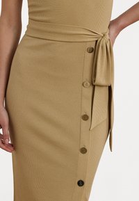 Khaki ribbad midiklänning med sidoknytning, som har framträdande knappar längs framsidan och en snug passform. Mjuk textur, avslappnad design.