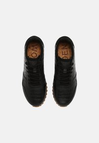 Paire de baskets en cuir noir texturé avec des lacets noirs et des semelles rainurées marron, vues de dessus sur un fond clair.