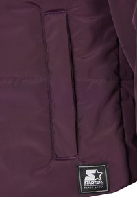 Gros plan d'une poche de veste matelassée violette avec une ouverture rectangulaire cousue et un patch logo noir "Starter Black Label".