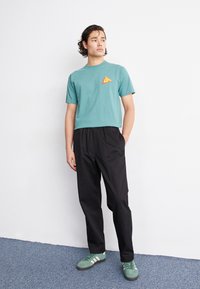 Camiseta de algodón color azul verdoso con un pequeño gráfico triangular naranja, combinada con pantalones negros de corte holgado y zapatos verdes a rayas.