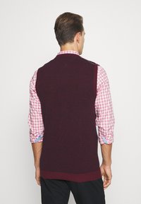 Bordeauxrode gebreide spencer met een gestructureerd patroon, gecombineerd met een roze geruit overhemd. Het overhemd heeft opgerolde mouwen en blauwe accenten.