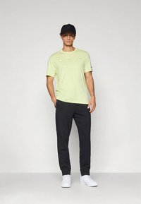 Light green cotton t-shirt with "Champion" logo, paired with dark gray sweatpants and white sneakers; model wears a black cap.
