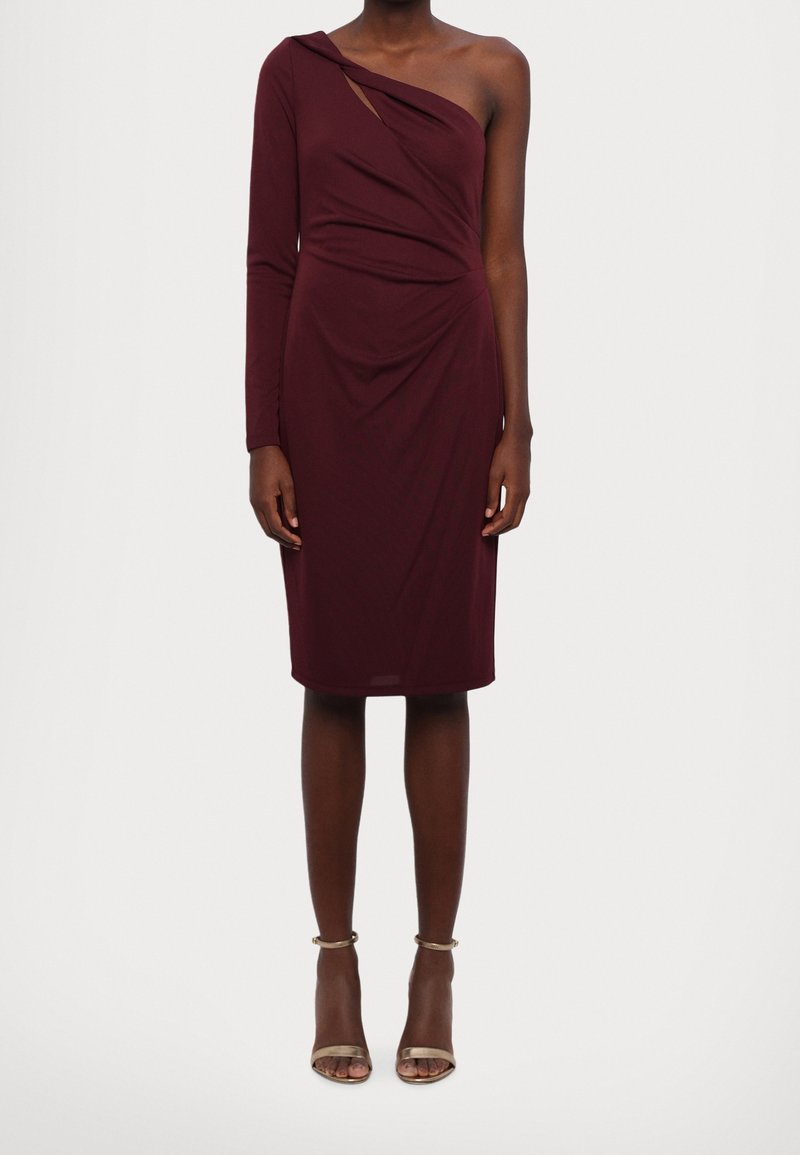 Kvinna i en kroppsnära, knälång bordeaux klänning med en lång ärm och en bar axel, matchad med metalliska ankelbandsskor.