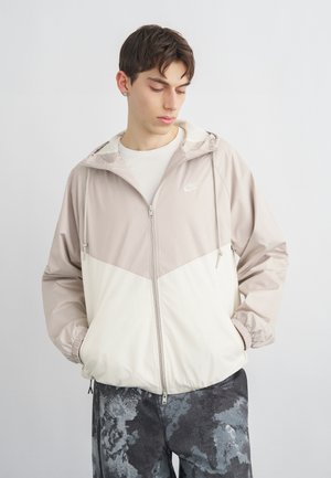 Νεαρός άνδρας που φοράει μπεζ και λευκό μπουφάν με φερμουάρ Nike, κοιτάζοντας κάτω με τα χέρια στις τσέπες, μπροστά σε απλό ανοιχτό φόντο.