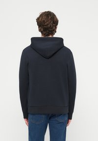 Tmavomodrá mikina s kapucňou s rebrovaným lemom a manžetami, s hladkou textúrou a minimalistickým dizajnom. Nosená s modrými džínsami.