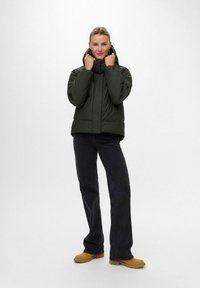 Chaqueta de plumas de color verde oscuro con cuello alto, combinada con jeans de pierna ancha en negro y botas de color beige. Textura suave, diseño versátil.