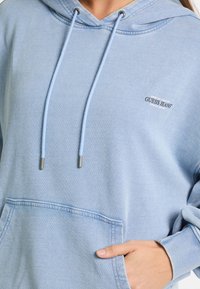 Gaiši zils kapuci krekls, kas izgatavots no kokvilnas maisījuma, ar priekšējo kabatu, regulējamiem aukliņām un izšūtu logo uz kreisā krūšu daļa.