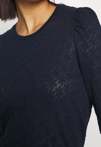 Tmavě modrý pletený top s texturovaným vzorem a jemnou průhledností. Má kulatý výstřih a nafouklé krátké rukávy.