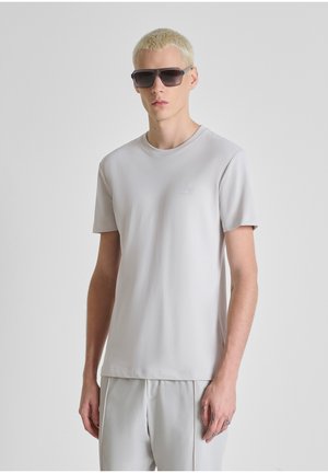 Hombre joven con cabello rubio corto, lleva gafas de sol grandes y oscuras, camiseta gris claro y pantalones holgados a juego, sobre un fondo sencillo.