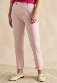 Gaiši rozā kokvilnas bikses ar pielāgotu griezumu, uzlocītām piedurknēm un pogu aizdari, kopā ar krēmkrāsas loafers kurpes ar zelta akcentiem.