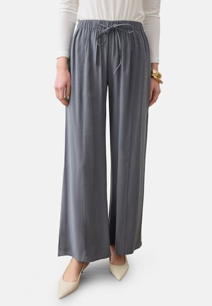 Vrouw draagt losse grijze wijde broek met koordsluiting in de taille, wit shirt met lange mouwen, gouden armbanden en beige puntige schoenen.