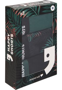 Tre paia di pantaloni corti in una scatola. Caratterizzati da colori verde scuro e nero con un motivo a foglie di palma. La scatola presenta un design tropicale verde e branding.