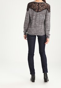 Maglione grigio con schiena in pizzo nero, maniche lunghe e tessuto testurizzato, abbinato a jeans blu scuro e stivali neri alla caviglia.
