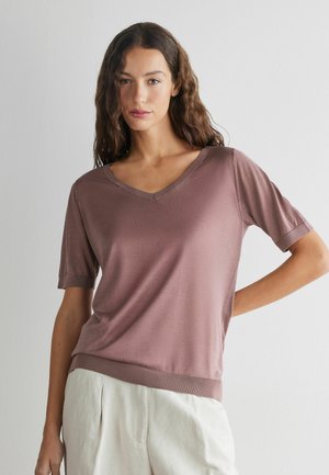 Kvinde med langt bølget hår iført en kort ærmet top med V-hals i farven mauve og lyse beige bukser, stående mod en ensfarvet lys væg.