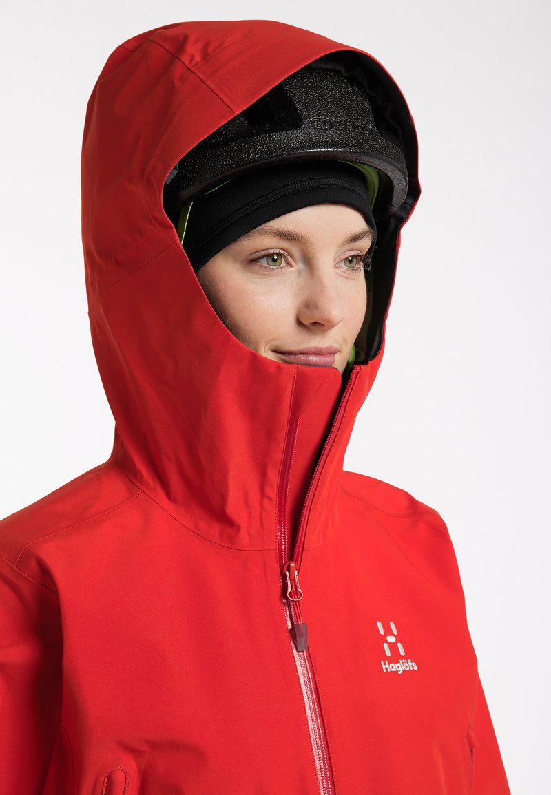 Osoba v červené bundě s kapucí, pod kterou má černou helmu a kuklu, s mírným úsměvem se dívá doprava na pozadí čistě bílé.