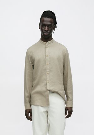 Mann mit dunkler Haut und Dreadlocks, der ein hellbeiges Hemd mit Stehkragen und weiße Hose trägt und vor einem schlichten hellen Hintergrund steht.