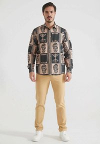 Chemise à manches longues dans un motif géométrique beige et noir, dotée d'un col classique et d'une fermeture à boutons à l'avant, associée à un pantalon beige et des baskets blanches.