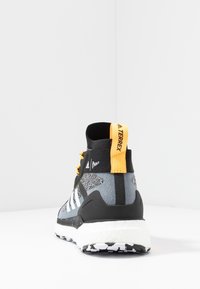 Baskets montantes noires et grises avec des rayures blanches, des accents jaunes, et une tige en tissu texturé. Elles présentent une semelle blanche épaisse et une languette de tirage.