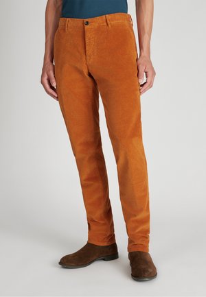 Orange Cord-Hosen mit schmalem Schnitt, vertikaler Rippung, Knopfverschluss und Seitentaschen, ergänzt durch braune Knöchelschuhe.