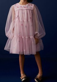 Flauschiges pinkes Tüllkleid mit bestickten Blumenmotiven, gestuftem Design und durchsichtigen Ballonärmeln. Silberne Schuhe runden das Outfit ab.
