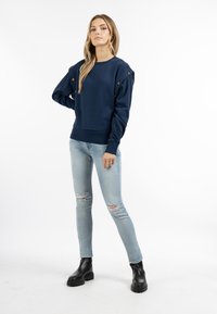 Felpa blu navy con bottoni sulle spalle, polsini e orlo a coste; abbinata a jeans strappati azzurri e stivaletti neri con suola spessa.