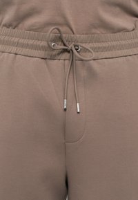 Lys brun joggebukse med elastisk liv med snøring, glatt tekstur og en sidelomme.