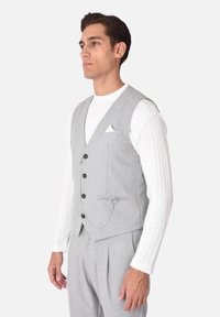Gilet grigio con quattro bottoni, una tasca frontale con un fazzoletto bianco, e un dettaglio a catena. Indossato sopra una camicia bianca strutturata.