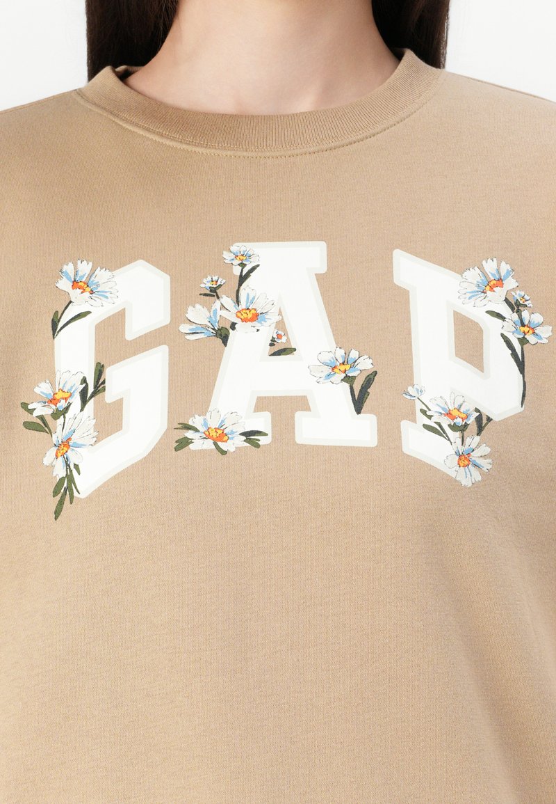 Sudadera beige con grandes letras blancas "GAP" decoradas con pequeñas flores blancas y azules en la parte superior del torso de una persona.