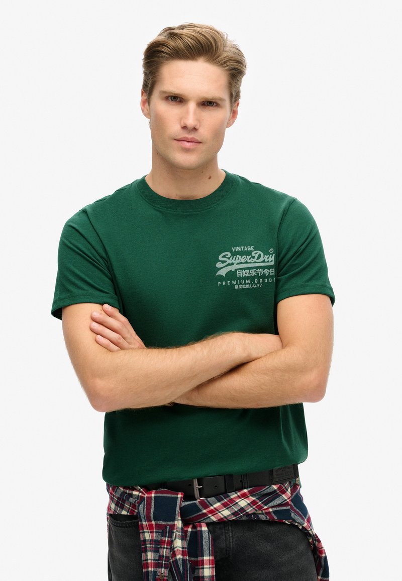 Grøn bomulds t-shirt med korte ærmer og rund hals; prydet med hvid vintage tekst på brystet; parret med en ternet skjorte bundet om livet.