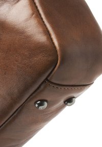 Eck einer braunen Ledertasche mit sichtbaren Nähten, subtiler Textur und Metallnieten. Glatte Oberfläche mit einer leicht verwitterten Optik.