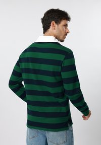 Άνδρας που φοράει πουκάμισο με μακριά μανίκια σε πράσινες και ναυτικό μπλε ρίγες με λευκό γιακά, στραμμένος πλαγίως, με ανοιχτό μπλε τζιν παντελόνι, πάνω σε απλό φόντο.