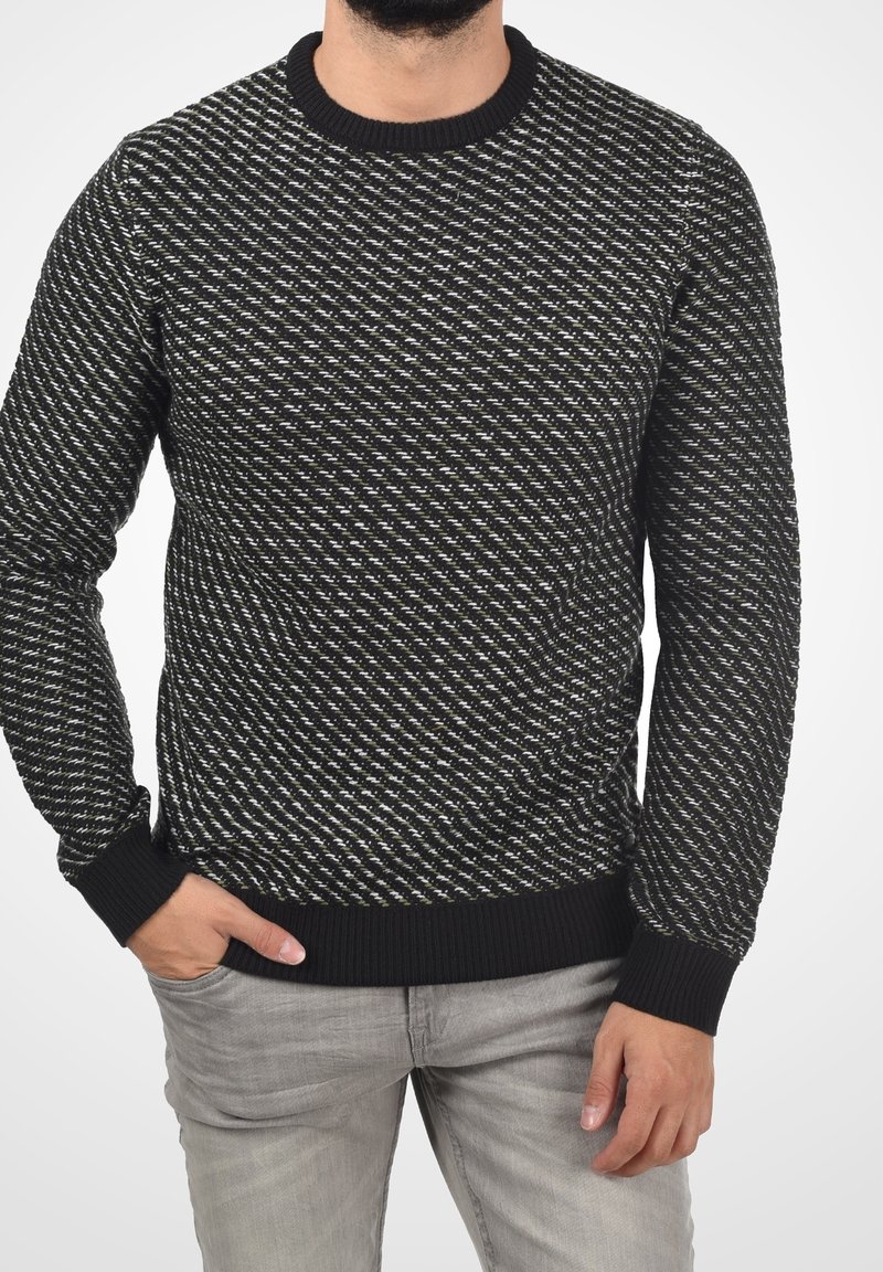 Uomo che indossa un maglione a girocollo a fantasia nera, bianca e verde oliva con polsini e orlo a coste, abbinato a jeans grigio chiaro.