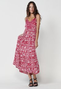 Rotes Maxikleid mit weißen, filigranen Mustern, Spaghetti-Trägern und einem gestuften Saum. Getragen mit schwarzen Riemchen-Sandalen.